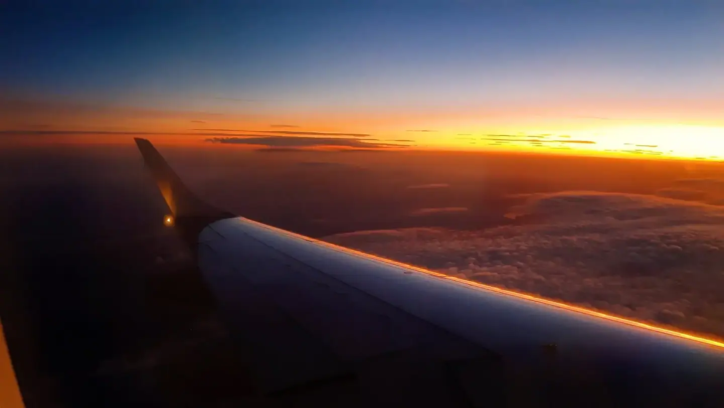 View of an airplane wing at sunset, flying above clouds with glowing orange and yellow horizon and a darkening blue sky.