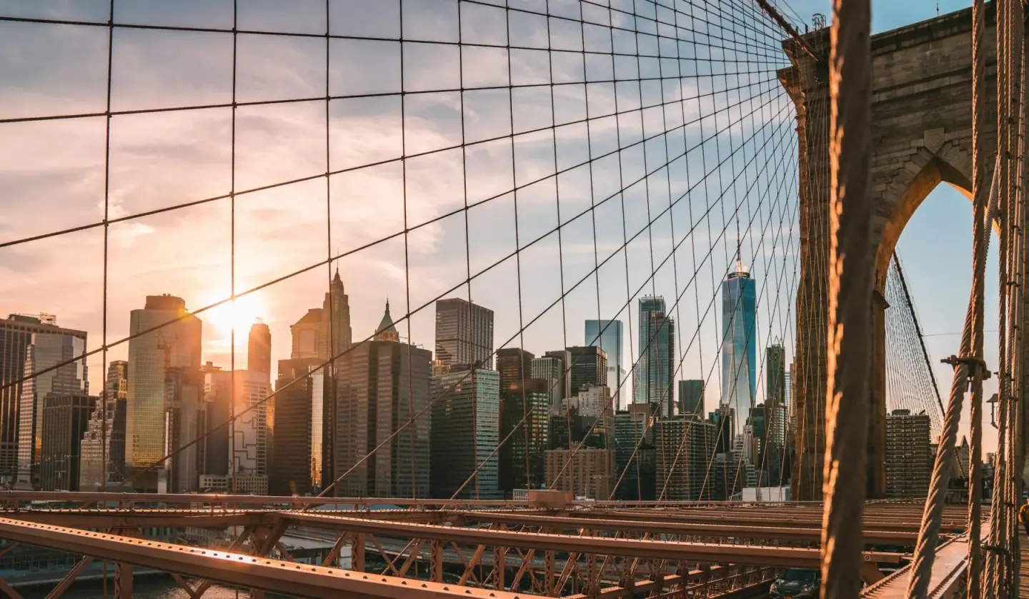 View of New York City skyline at sunset through the Brooklyn Bridge cables.